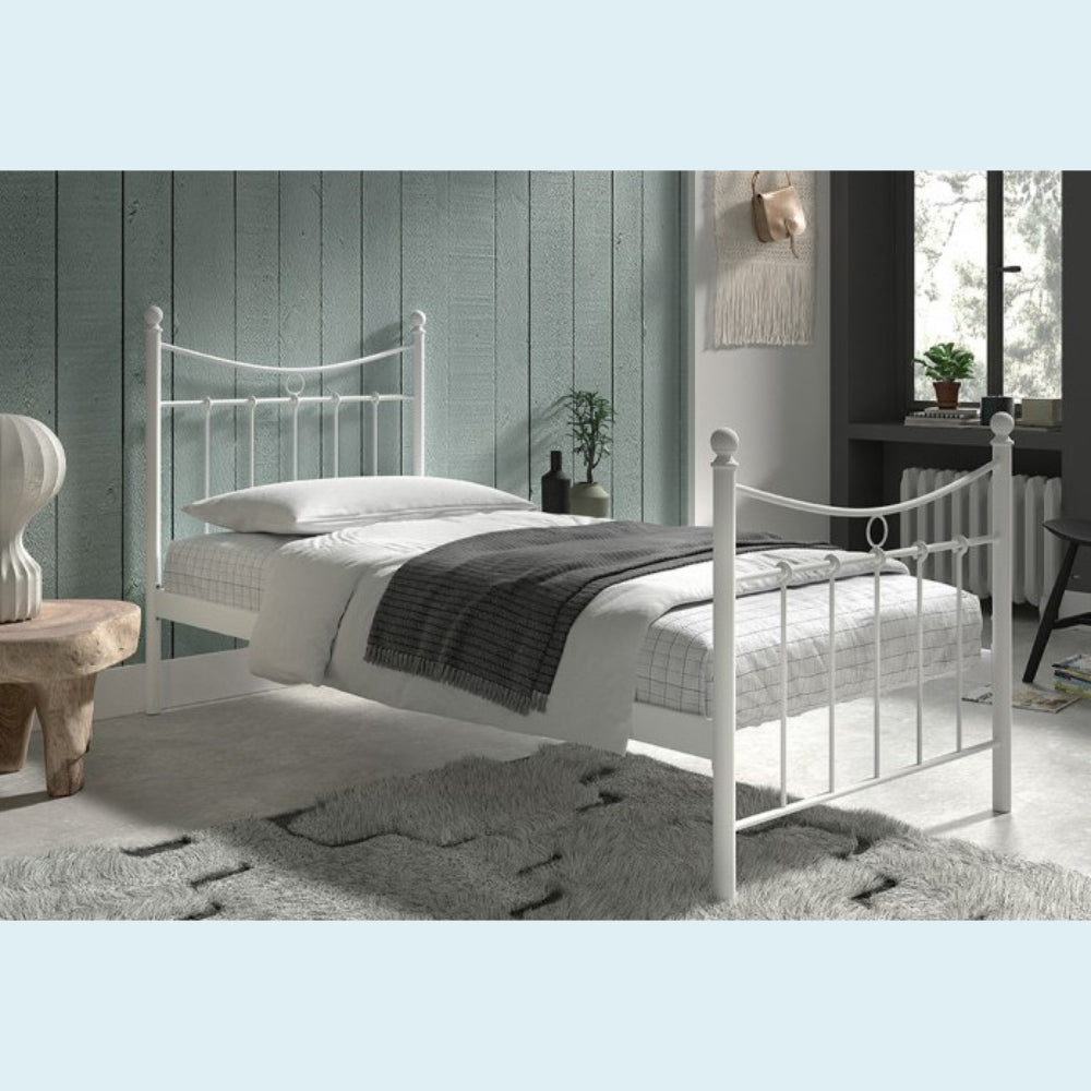 Wit metalen bedframe met wit beddengoed en een grijze plaid, geplaatst in een gezellige kamer met een groene muur en een groot raam.