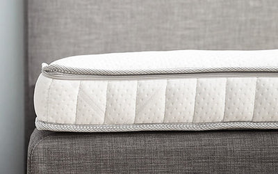 Close-up van een wit matras met grijze biezen, geplaatst op een grijs gestoffeerd bedframe.
