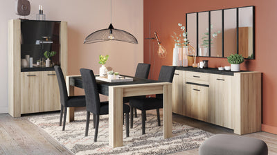 Moderne eetkamer met een houten eettafel en zwarte stoelen, een vitrinekast en dressoir in een lichte houtkleur met zwarte accenten, decoratieve accessoires en een grote hanglamp, tegen een terracotta muur.